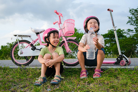 小女孩喝水摄影照片_快乐可爱的小女孩和她的妹妹坐在路边的草坪上，喝着公园里自行车附近的瓶子里的水。