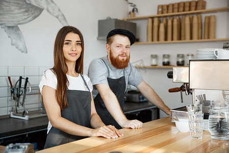 咖啡经营理念-积极的年轻留着胡子的男人和美丽迷人的女咖啡师夫妇穿着围裙，站在酒吧库特准备在现代咖啡店提供咖啡服务