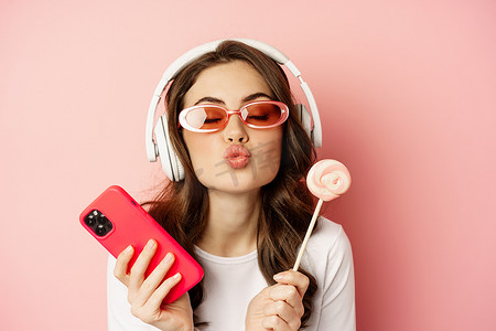 漂亮的女模特戴着耳机听音乐，拿着棒棒糖和手机，戴着太阳镜，站在粉色背景上