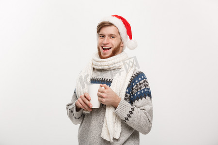 圣诞节概念 — 穿着毛衣和圣诞老人​​帽子的年轻胡子男子拿着一个热咖啡杯，用白色隔开，上面有复制空间。