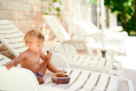 小女孩坐在阴凉处的躺椅上，从水果盒里取出草莓
