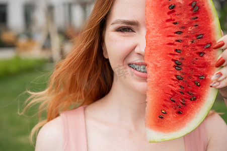 张牙舞爪狮子摄影照片_夏天，美丽的红发女人戴着牙套微笑，用一片西瓜遮住半张脸