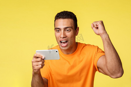 身穿橙色 T 恤、欢快兴奋的年轻帅哥举起拳头，为最喜欢的球队加油，水平拿着智能手机，在移动设备上看足球，黄色背景