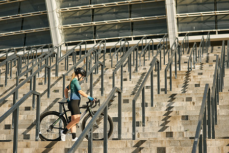 女自行车运动员摄影照片_年轻的职业女自行车手在户外锻炼时，穿着自行车服和防护装备，骑着自行车站在台阶上