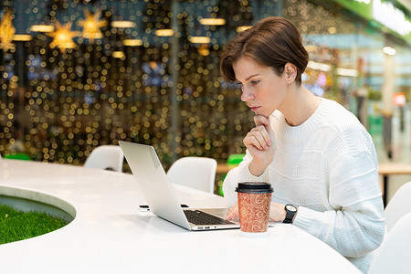 照片中，一位年轻女子坐在购物中心美食广场的一张桌子旁，在笔记本电脑前工作。