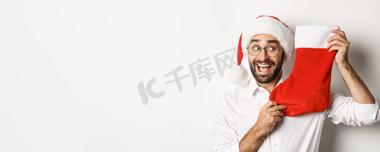 圣诞袜里有礼物摄影照片_快乐男人庆祝圣诞节的特写，收到圣诞袜里的礼物，看上去很兴奋，戴着圣诞帽和眼镜，白色背景