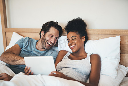 快乐、轻松、无忧无虑的夫妻在数字平板电脑上阅读社交媒体新闻，躺在床上大笑。
