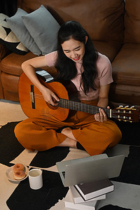 劳动节快乐字体摄影照片_快乐的女人学习弹吉他，在舒适的客厅地毯上使用笔记本电脑