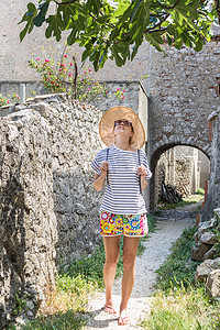 美丽的金发年轻女旅行者戴着草帽，在地中海享受夏天，​​在一棵无花果树下采摘水果，树上有薰衣草花，背景是传统的老石屋