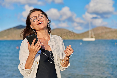 眼睛闭摄影照片_戴着耳机在海滩上用智能手机听音乐的快乐女人