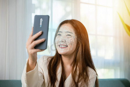 亚洲女性使用智能手机进行门禁人脸识别，在家中沙发客厅进行私人身份识别