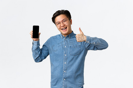 乐观而满意的亚洲男子戴着牙套，戴着眼镜，穿着休闲服，竖起大拇指表示批准智能手机应用程序，面对手机屏幕推广应用程序，白色背景