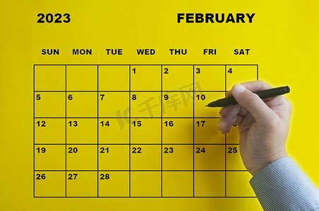 2023 年 2 月日历，黄色背景上手持黑笔。