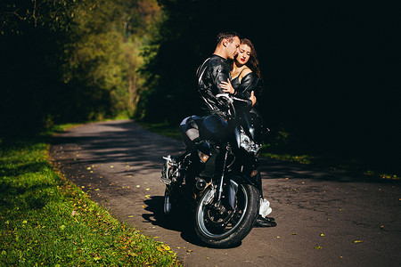 英俊的年轻人和美丽的年轻女孩，夫妇坐在摩托车上，站在一辆穿着黑色皮衣的摩托车附近，拥抱，在大自然中，在街上