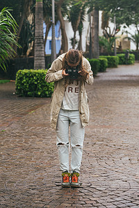 反光图片摄影照片_年轻的时髦女性拿着反光相机，在湿石路面上拍摄靴子的照片
