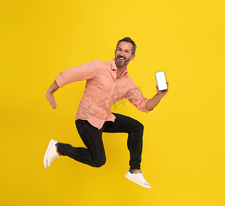 智能手机手机摄影照片_成熟的灰发男子跳高，手里拿着智能手机，在镜头前开心地微笑着，身穿桃色衬衫和黑色牛仔裤，背景为黄色。