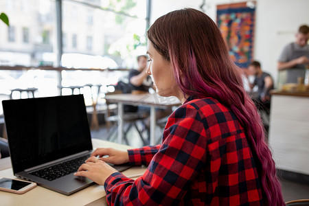 一位年轻的粉红色头发女性坐在咖啡馆里，在带有空白复制空间屏幕的笔记本电脑上敲键盘的后视图。