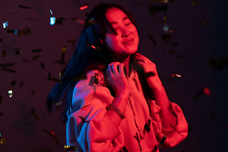 庆祝霓虹灯摄影照片_兴奋的亚洲美女跳舞，玩得很开心，为工作室里霓虹灯​​下的五彩纸屑雨而欢欣鼓舞。