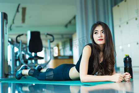 运动女子躺在有运动器材背景的健身馆的瑜伽垫上放松。