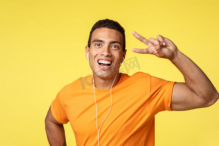 巴西音乐摄影照片_穿着橙色 T 恤的快乐、幸运的微笑男人，戴着耳机听励志音乐，展示和平或胜利的标志，咧嘴笑着乐观，欣赏最喜欢的歌曲，站在黄色背景中乐观
