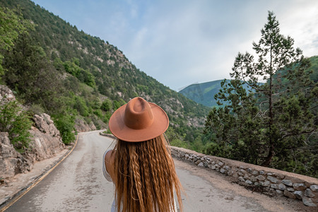 穿着棕色帽子和白色连衣裙的女旅行者看着令人惊叹的山脉和森林、流浪旅游概念、大气的史诗时刻