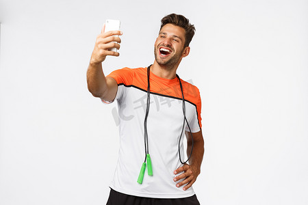 英俊的男运动员在健身房训练后自拍，手持智能手机，伸出手臂，满意而自豪地微笑，告诉追随者关于跳绳、锻炼和互联网概念的新记录