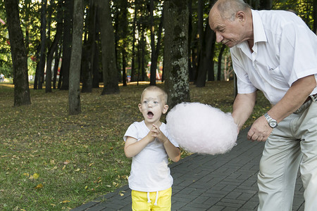 对待孩子摄影照片_在游乐园里，祖父慈爱地、细心地对待他的小孙子，并给他喂粉红色的棉花糖。