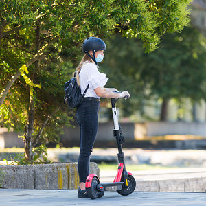 时尚时尚的少女在公共场合戴着电晕病毒防护面罩，同时在城市环境中使用租赁电动滑板车。