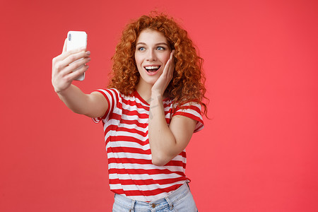 时尚可爱的红发卷发女性夏季 T 恤录制视频社交媒体，来自豪华热带度假村出国旅游度假自拍手持智能手机摆出愚蠢的照片红色背景