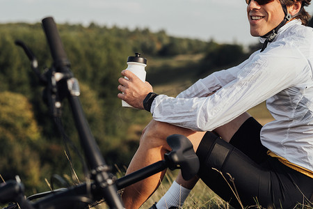 职业男性骑自行车的人从瓶子里喝水，男人在乡下户外旅行时坐在自行车旁