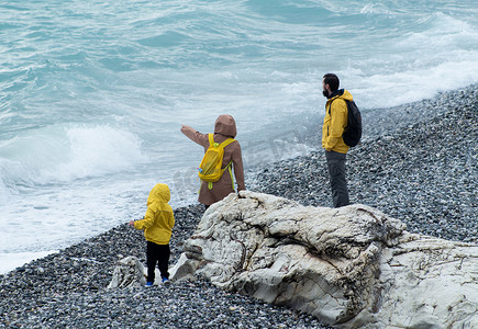 2022岩石摄影照片_俄罗斯索契 — 2022 年 5 月 9 日：一个男人、一个女人和一个孩子穿着黄色雨衣沿着鹅卵石海滩散步