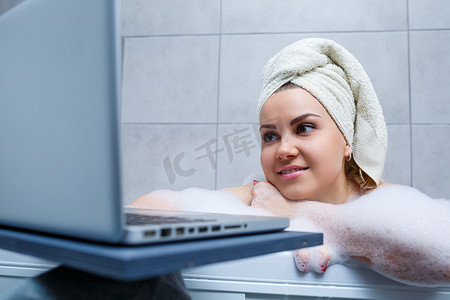 一位头戴毛巾的年轻女子坐在美容院的浴缸里，用笔记本电脑看电影。