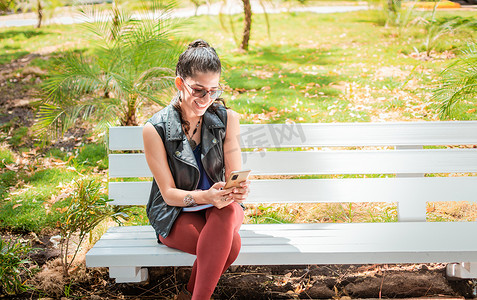 女孩坐在长椅上看手机，快乐的女人坐在公园里用手机发短信，女人坐在长椅上发短信