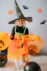 一个女巫摄影照片_儿童万圣节-一个戴着女巫帽的女孩和一个狂欢节服装，家里有通风的橙色和黑色气球。