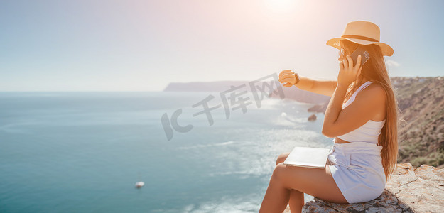 坐在海边的女摄影照片_数字游牧者，戴帽子的女人，一位带着笔记本电脑的女商人在日落时坐在海边的岩石上，从远处进行在线商业交易。