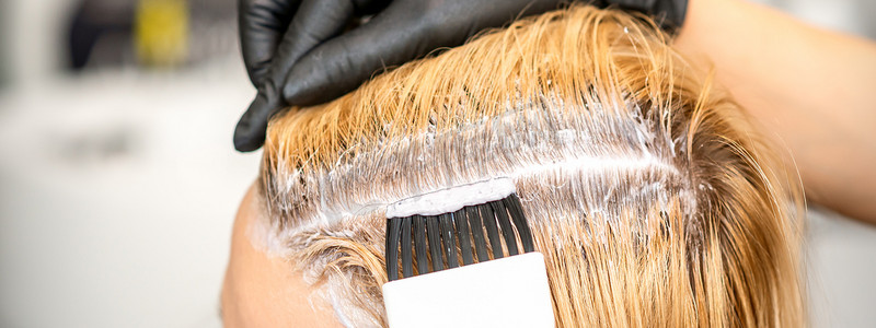 染头摄影照片_美发师正在美发沙龙里用刷子为一位年轻女子染金色的发根。