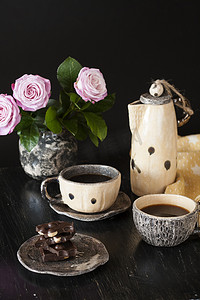 两杯黑咖啡、黑巧克力、一个黄色茶壶和一个深色背景上插着粉红玫瑰的花瓶。