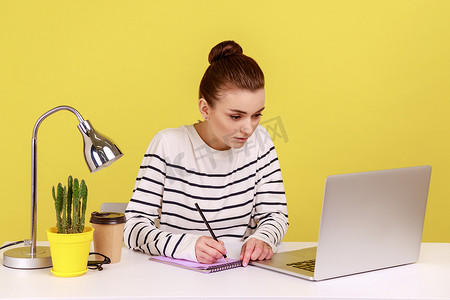 集中精力的女上班族用铅笔在记事本上做笔记，看着笔记本电脑显示屏。