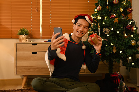 戴着圣诞老人帽子的微笑男子在家里的圣诞树附近用智能手机自拍。