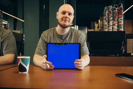 快乐的男性坐在现代咖啡馆附近的长凳上，用手指指着平板电脑的空蓝屏