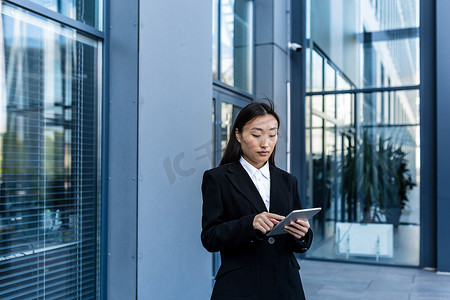 商务正装摄影照片_身穿商务正装的亚洲女商人站着，手里拿着平板电脑工作