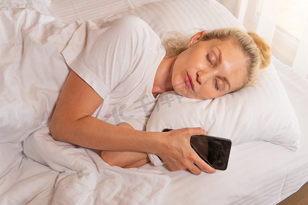 女人美丽的手机金发女郎睡眠移动床毯躺在床上，从成人枕头做梦和家庭小睡，睡前灯。