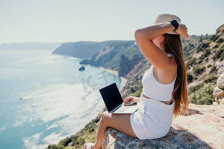 数字游牧者，戴帽子的女人，一位带着笔记本电脑的女商人在日落时坐在海边的岩石上，从远处进行在线商业交易。