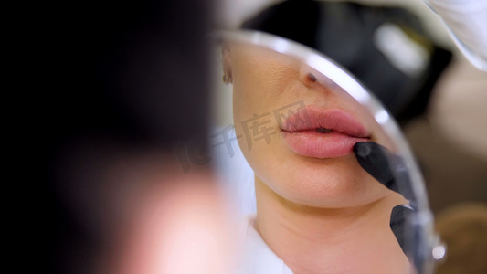 医疗办公室，美丽的女人照镜子，医生在嘴唇注射透明质酸后探测病人的嘴唇，矫正嘴唇的形状。