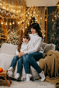 家庭环境摄影照片_一个小女孩和她的母亲在舒适的家庭环境中，坐在圣诞树旁边的沙发上。