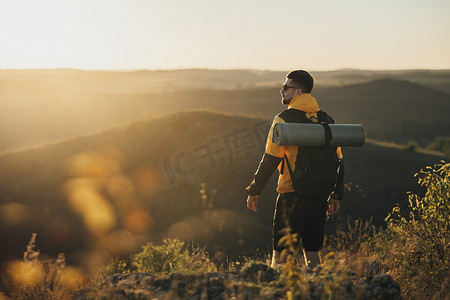 后视图摄影照片_一名背着背包和野营垫的旅行男子的后视图，日落时站在山顶上，男性游牧者享受他的独行旅行