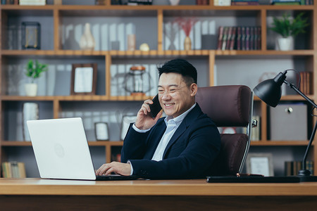 行政文员摄影照片_快乐的亚洲银行家老板在经典办公室里用笔记本电脑打电话