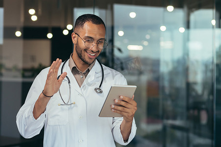穿着医用白大褂和听诊器的快乐的西班牙裔医生使用平板电脑为患者进行视频通话和在线咨询，在诊所办公楼内工作的男子