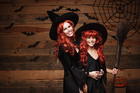美丽的白人母亲和她的女儿，长着红色的长发，穿着巫婆服装庆祝万圣节，在木制工作室背景上摆出蝙蝠和蜘蛛网的姿势。