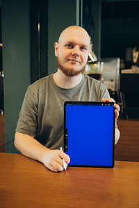 app蓝摄影照片_坐在现代咖啡馆附近的长椅上，快乐的男性用手指指着空白的平板电脑蓝屏